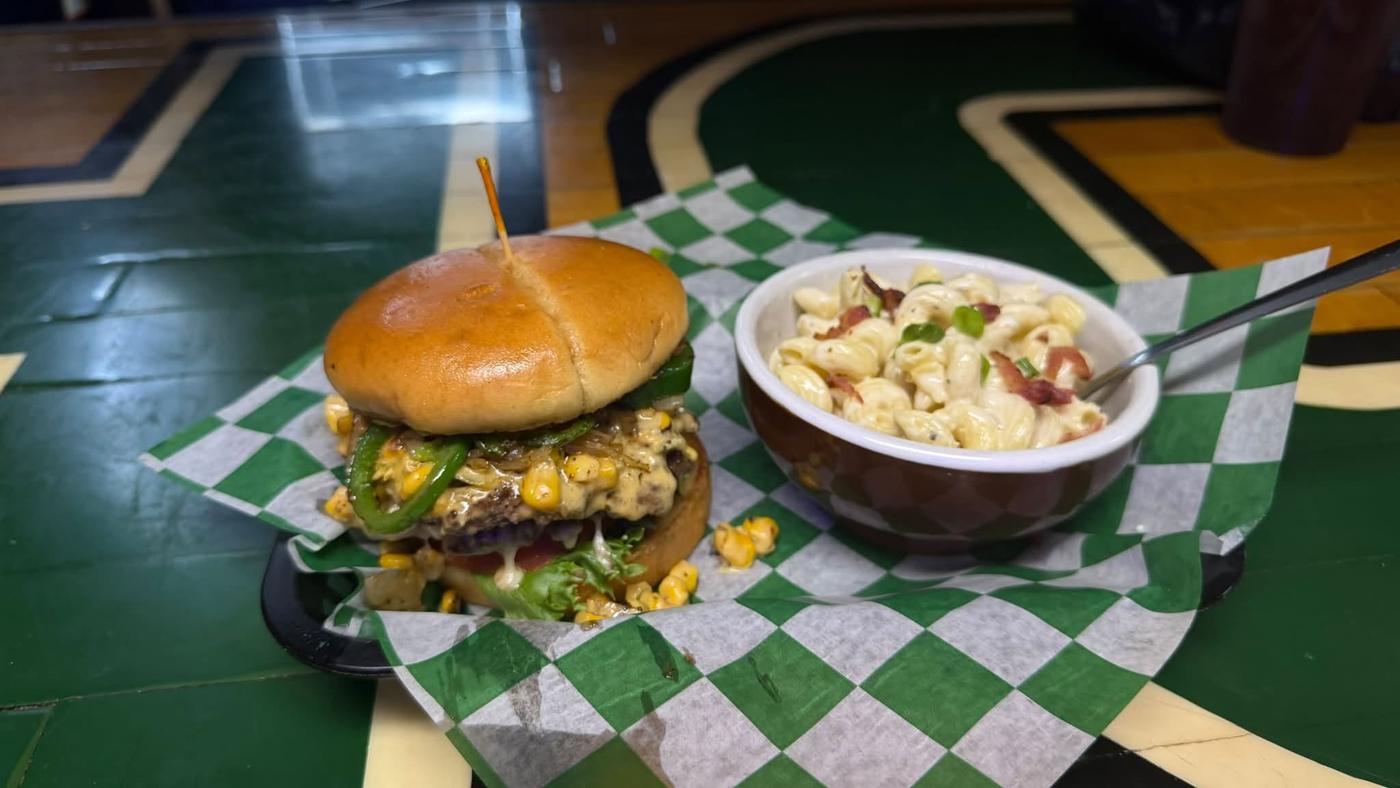A burger topped with grilled peppers and corn relish beside a bowl of bacon mac and cheese, on green checked paper