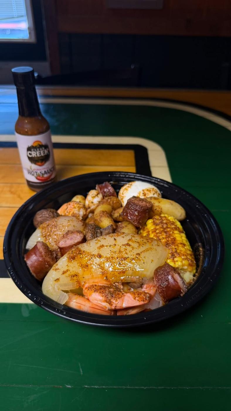 A shrimp boil with sausage, corn, potatoes and onion in a takeout bowl, with a bottle of Clear Creek hot sauce behind it